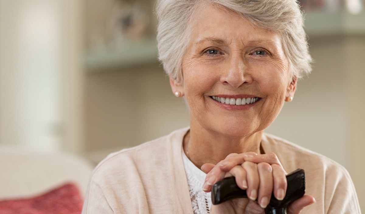 Freundlich lächelnde, ältere Dame mit einem Gehstock in der Hand, sitzend, in häuslichem Umfeld